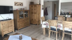 A living room with a TV, wooden cabinets, a dining table with chairs, and a kitchen area in the background. - Nendaz Centre Superbe Appartement 3 p. 75m2 Avec Piscine Parking et vue (Nendaz)