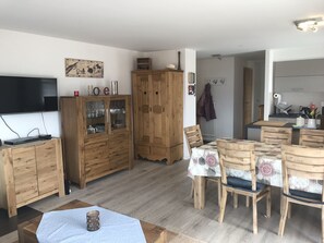 Ein Wohnzimmer mit Fernseher, Holzschränken, einem Esstisch mit Stühlen und einer Küchenzeile im Hintergrund.