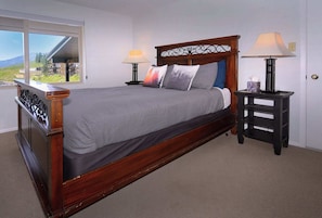 Un dormitorio con una cama de madera, dos mesitas de noche con lámparas, una ventana con vistas y una puerta.