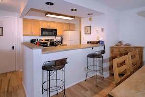 Una cocina compacta con una isla central, dos taburetes de bar, armarios de madera, un microondas y un refrigerador.