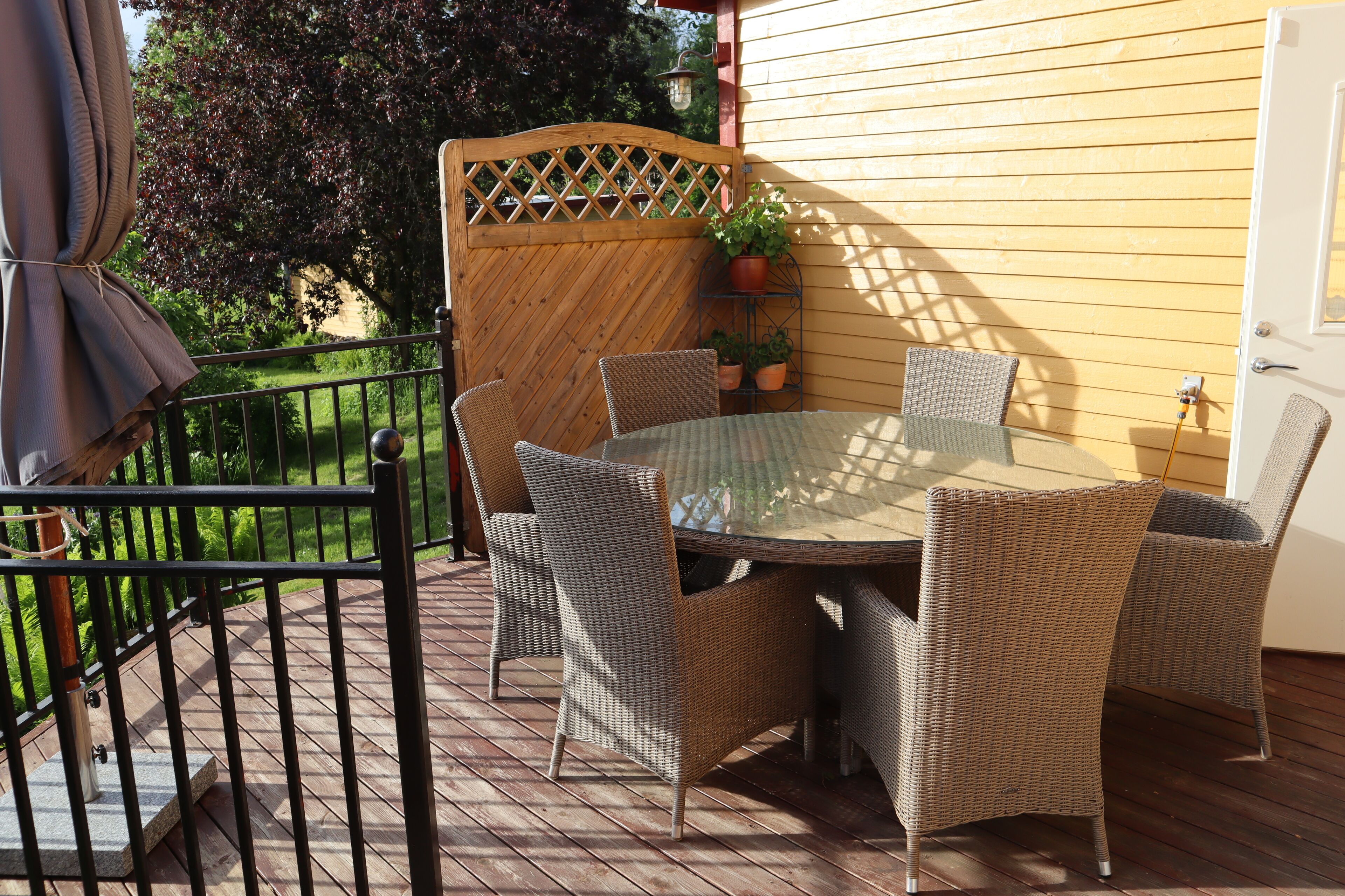 Une terrasse avec des chaises en osier et une table en verre, entourée d’une clôture en bois et d’un mur jaune.