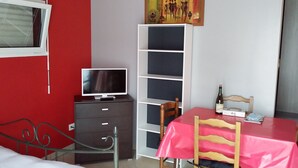 Una habitación pequeña y bien iluminada con una cama, un escritorio con una computadora, una silla, una mesa con mantel rojo y una estantería.