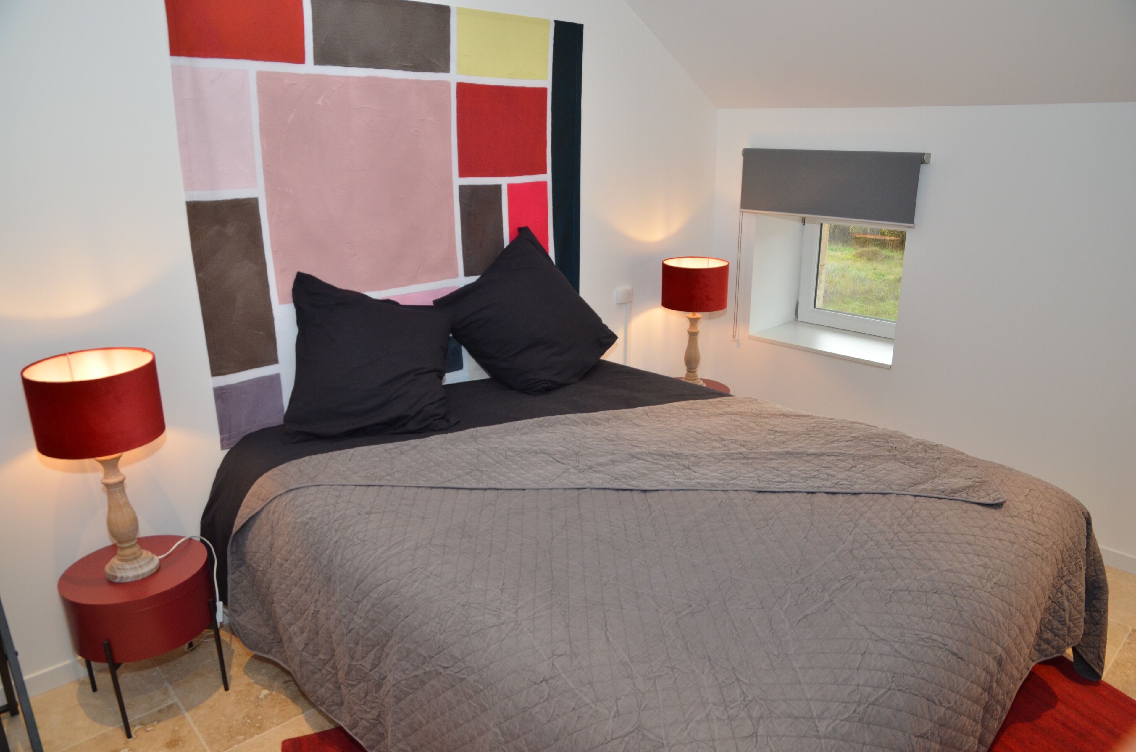 A bedroom with a large bed, two bedside lamps, a window with a blind, and a colorful wall art piece.