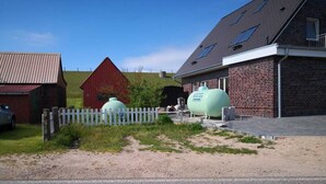 Maalaismaisema, jossa on punainen lato, valkoinen puinen aita ja kaksi suurta vihreää säiliötä, joissa on teksti ”Fischer Gas”, ja nämä sijaitsevat tiilisen rakennuksen lähellä.