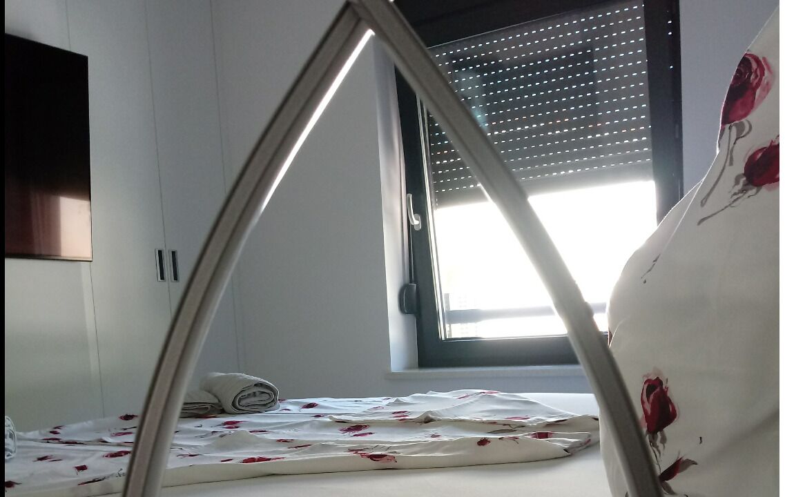A bedroom with a bed, a window with blinds, and a wall-mounted TV.