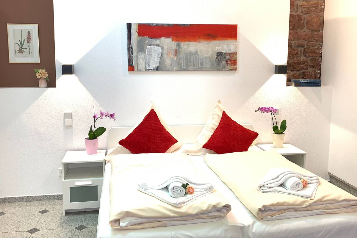 A hotel room with a bed, two red pillows, a nightstand with a pink vase and an orchid, a painting on the wall, and a brick wall on the right.