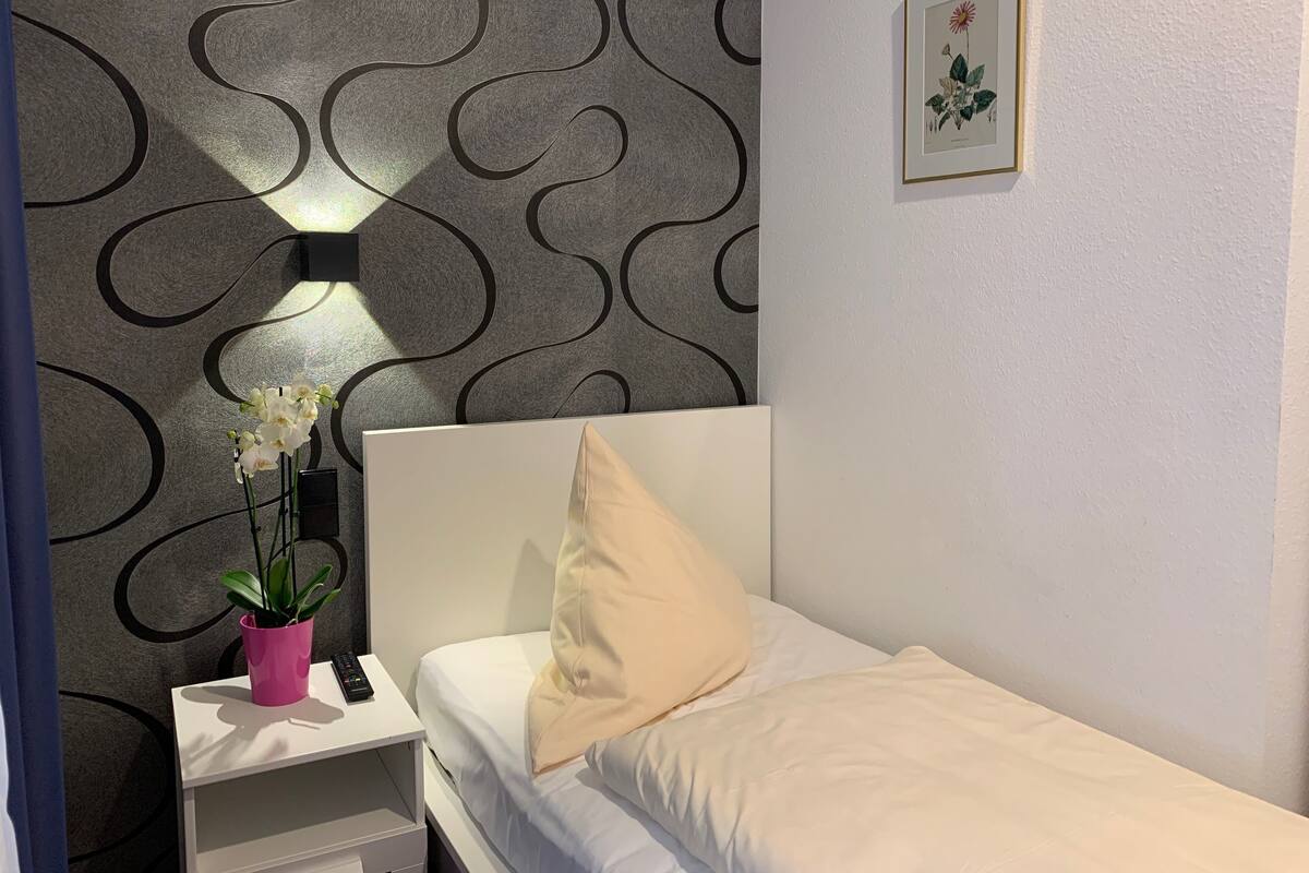 A neatly made bed with a white headboard and a single beige pillow, a small bedside table with a pink plant and a remote control, and a wall with a black and grey wavy pattern.