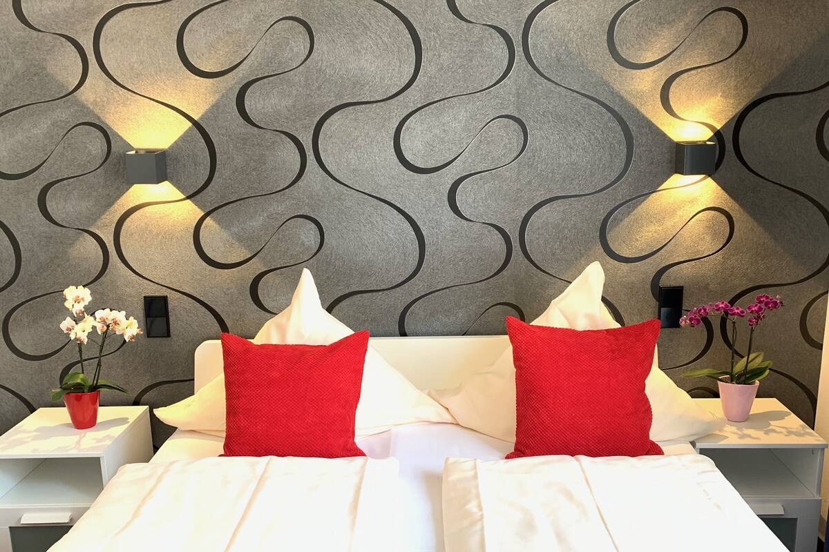 A hotel room with a bed, two red pillows, a white bedside table, a red vase with white flowers, and a wall with a wavy pattern.