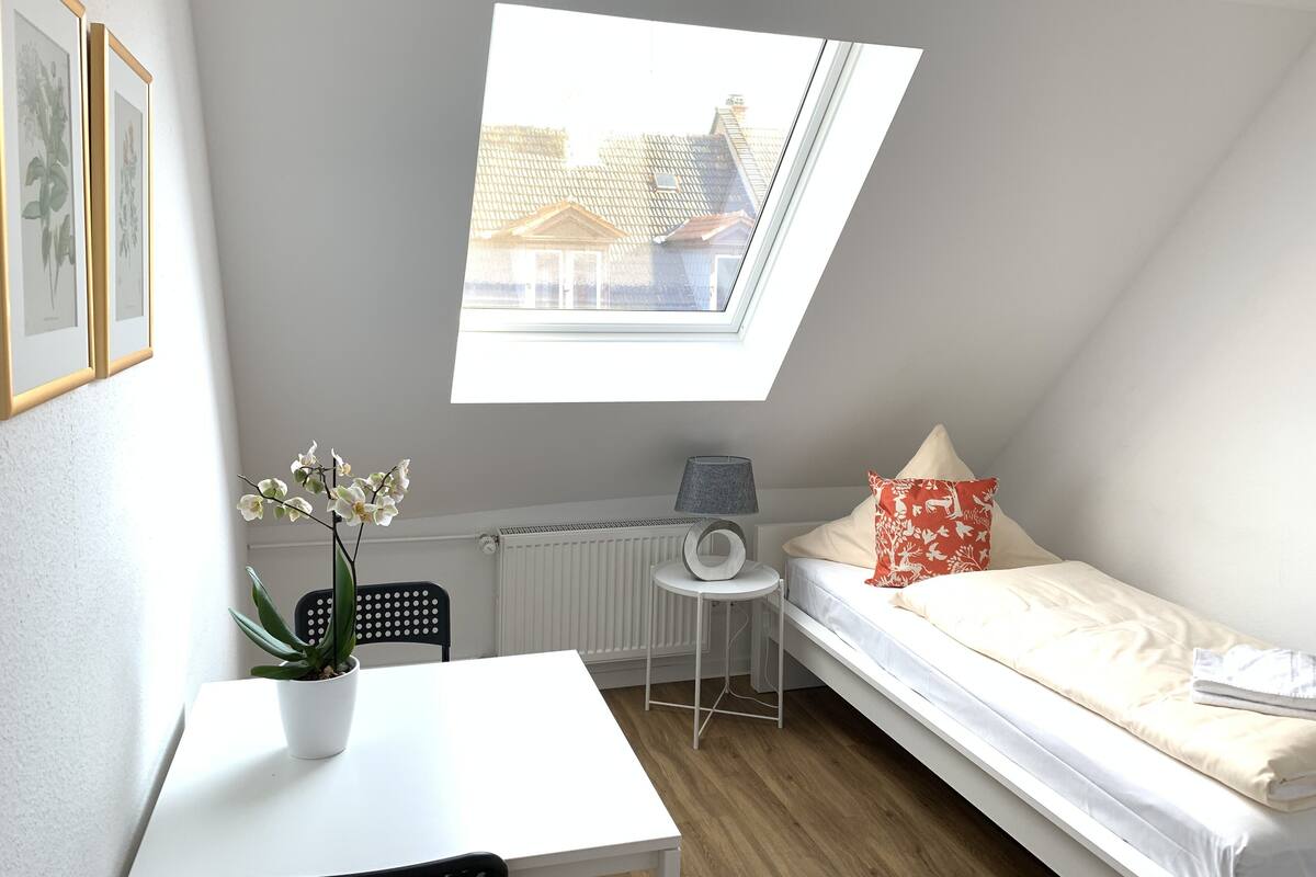 A small, bright room with a slanted ceiling, a single bed, a small table with a potted plant, and a radiator.