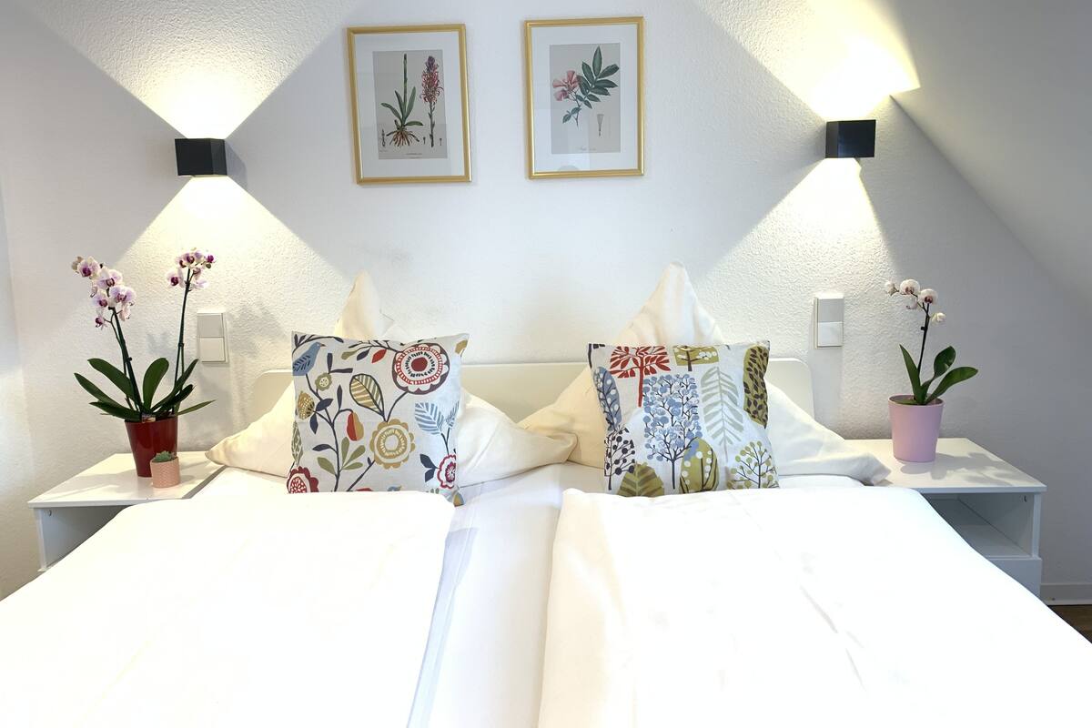 A hotel room with a bed, two nightstands, two wall-mounted lamps, two framed botanical prints, and two potted orchids.