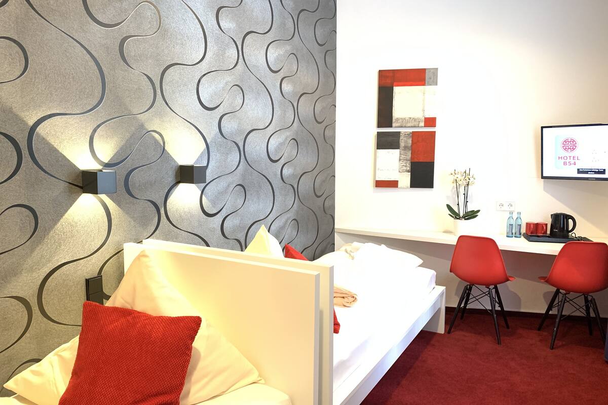 A hotel room with a modern design, featuring a bed with white linens and red pillows, a desk with two red chairs, and a wall-mounted TV displaying the hotel name.