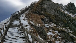 Eine Holztreppe führt einen einen felsigen Berghang mit Schneeflecken hinauf.