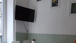 Ein kleiner, heller Raum mit einem weißen Tisch und vier Stühlen, einem an der Wand befestigten Fernseher und einer Heizung.