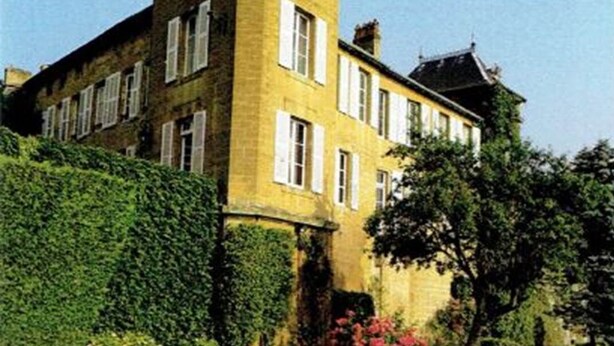 Un grand bâtiment historique, doté d’une tour imposante et de nombreuses fenêtres, entouré de jardins et de haies bien entretenus.