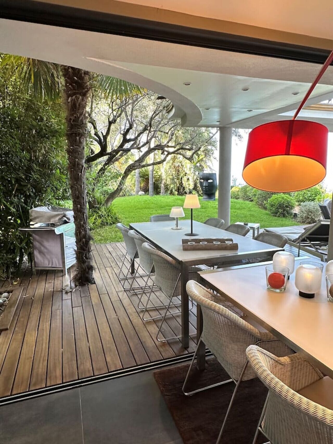 Un espace de restauration en plein air doté d’une terrasse en bois, d’une table avec des chaises, d’une suspension rouge et offrant une vue sur un jardin luxuriant.