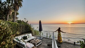 Une terrasse avec un canapé, une table et une personne vêtue d’une robe jaune, d’où l’on peut admirer l’océan au coucher du soleil.