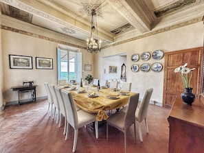 Een eetkamer met een grote tafel voor zes personen, omringd door stoelen, een kroonluchter en een uitzicht door een raam.