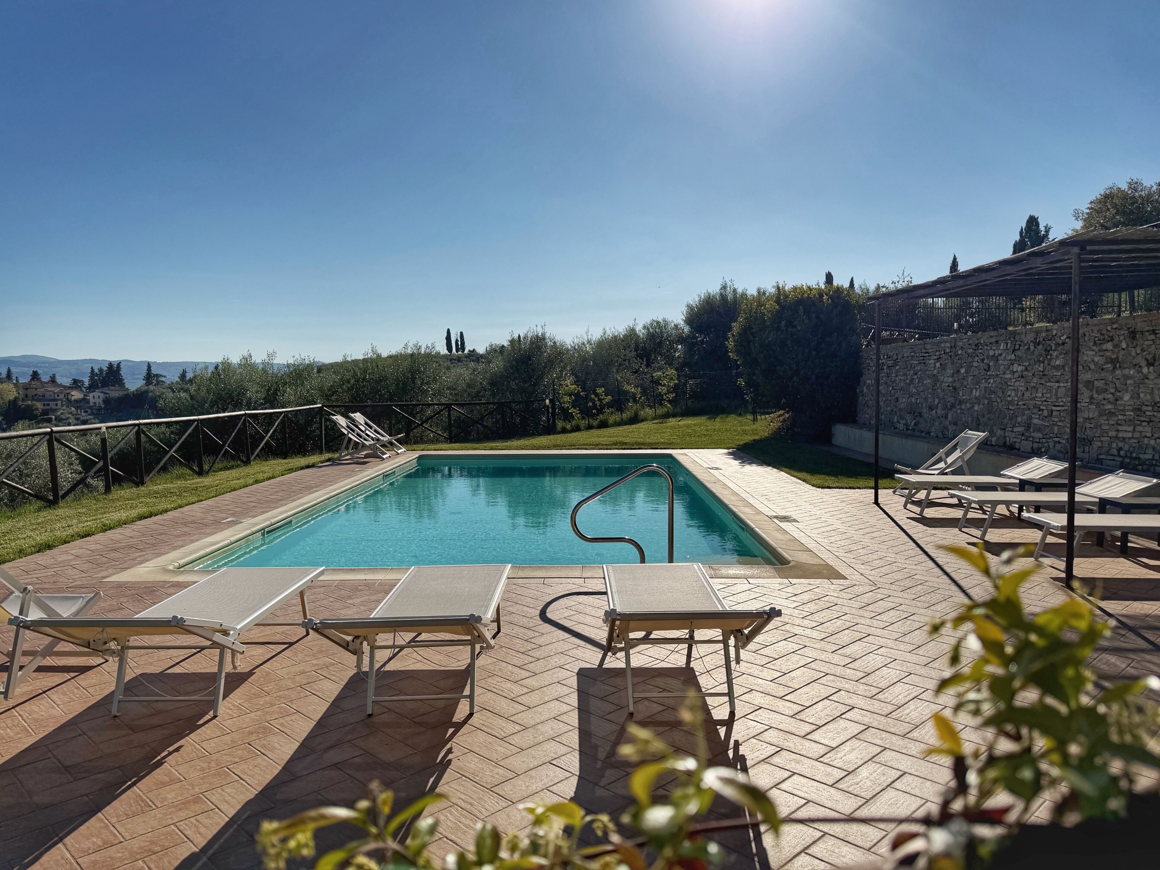 Una piscina rectangular con una escalera metálica, rodeada por un patio de ladrillo. Se observan sillones de descanso y un muro de piedra al fondo.