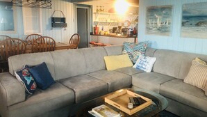 Een gezellige woonkamer met een grijze bank, een salontafel, en een eethoek met houten stoelen en een tafel.