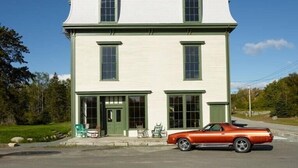 A two-story white building with green trim and a classic design, featuring a front porch with chairs and a vintage car parked outside. - Luxury Apartment overlooking Center Harbor in Brooklin (Brooklin)