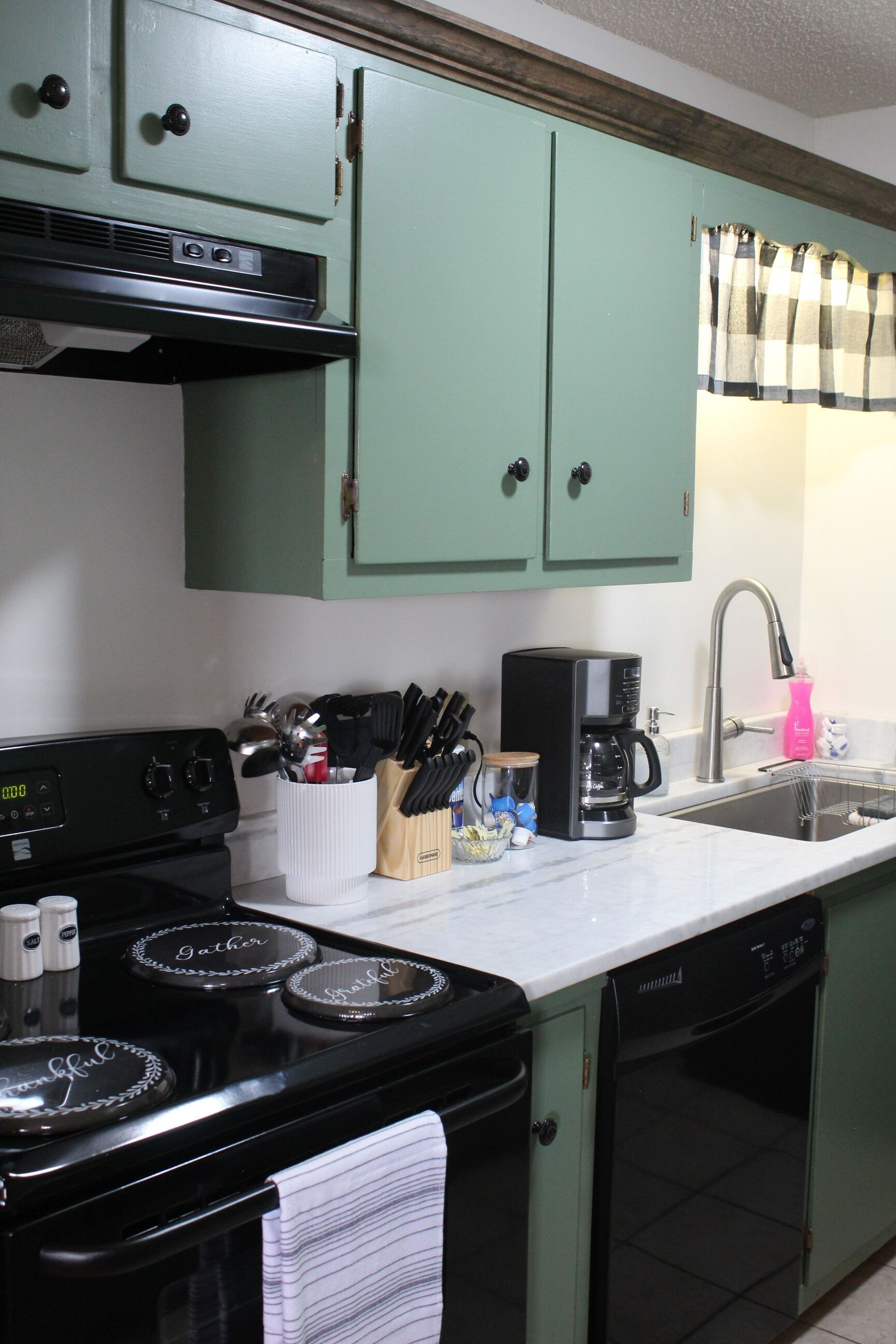 Une cuisine avec des meubles de couleur verte, une cuisinière noire, une machine à café et un lave-vaisselle.
