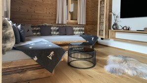 A cozy room with a wooden ceiling, a sofa with a fur throw, a round black coffee table, and a flat-screen TV mounted on the wall.