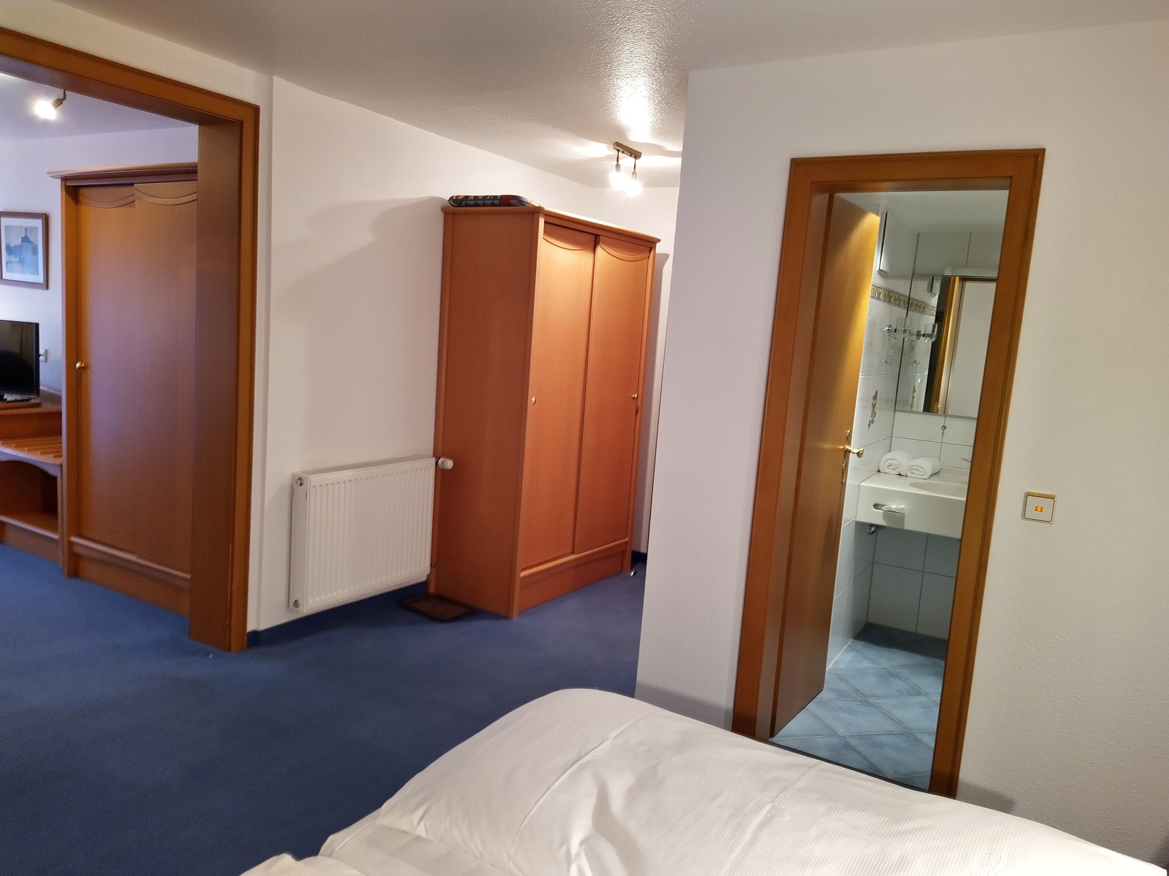 Une chambre à coucher avec un lit, une armoire, un radiateur et une salle de bain visible à travers une porte ouverte.