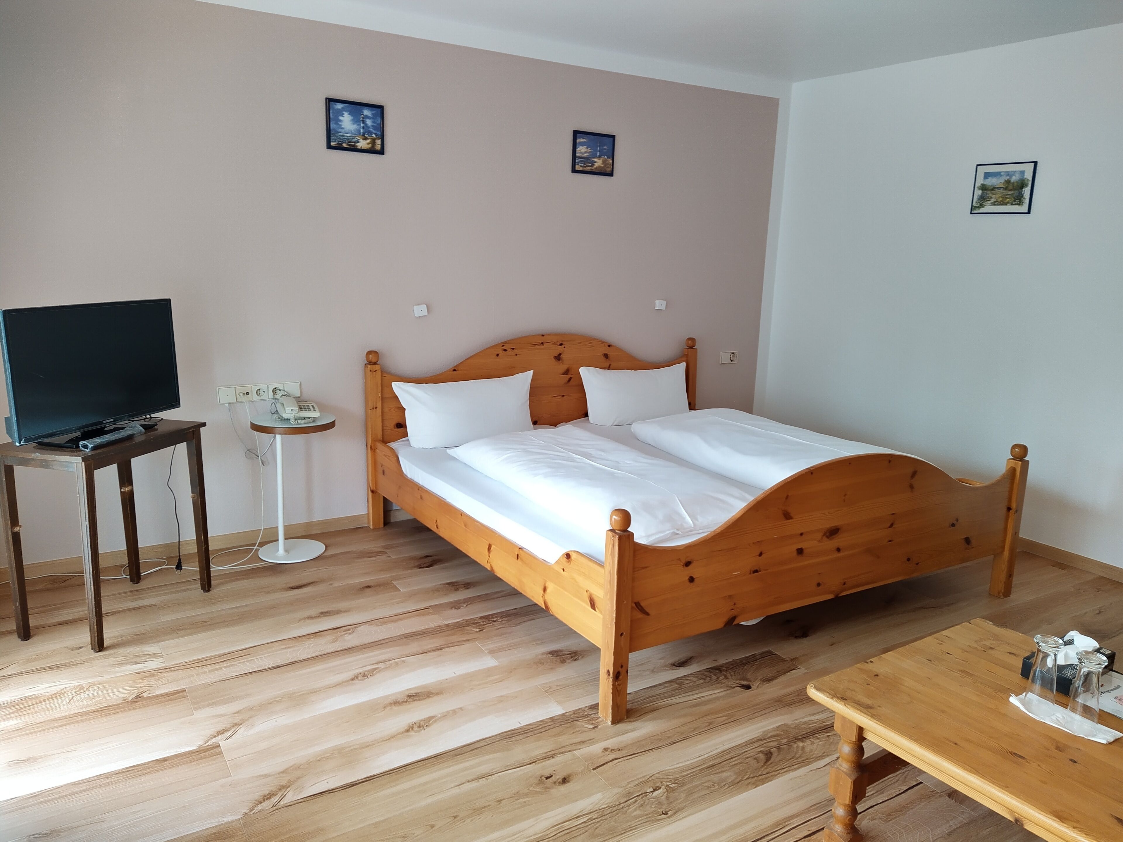 Un lit en bois avec du linge de lit blanc, un téléviseur posé sur un meuble, une petite table avec un téléphone et un cadre photo accroché au mur.