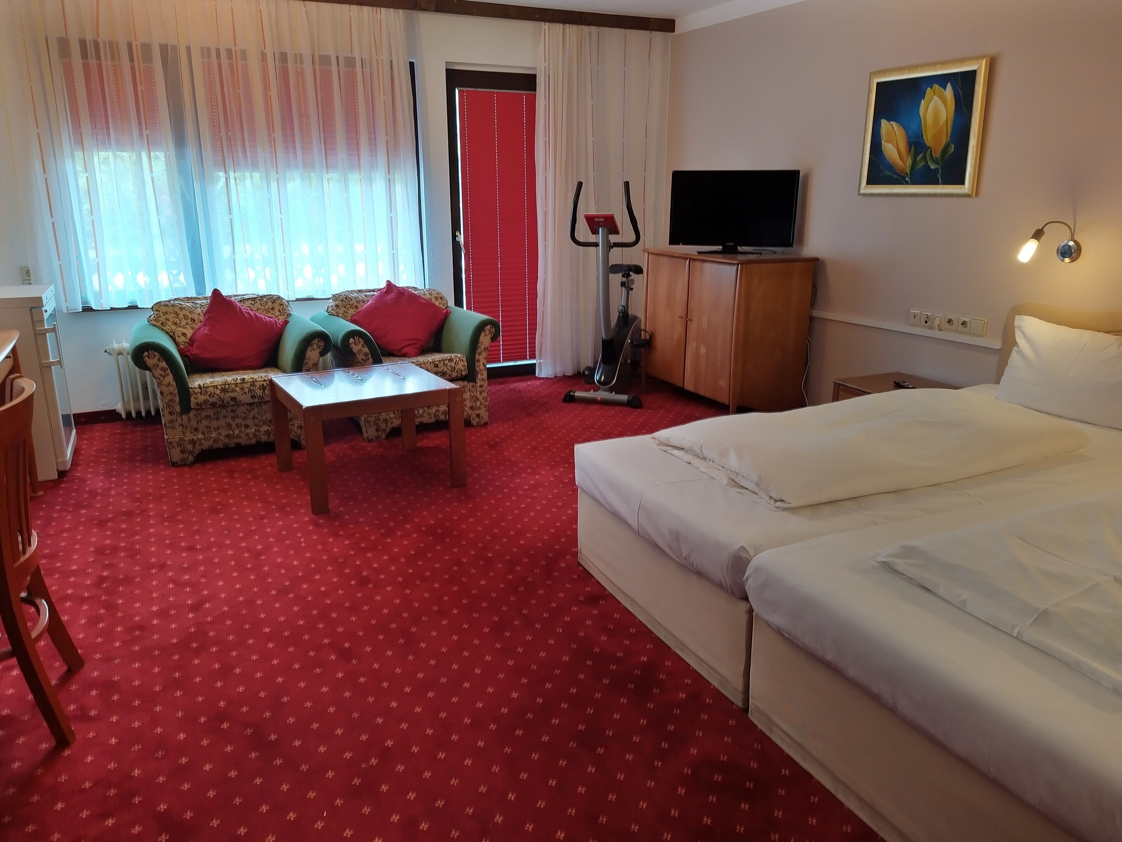 A hotel room with a red carpet, a sofa with floral upholstery, a wooden coffee table, a bed, a TV, and a painting on the wall.