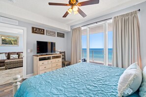 A bedroom with a bed, a TV, a dresser, a chair, and a balcony with a view of the ocean.