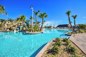 A swimming pool with a water slide, palm trees, and a thatched-roof building.