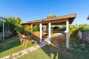 A covered outdoor seating area with wooden furniture and a stone pathway. - Palamutbükü Deniz Apart Otel (Datça)