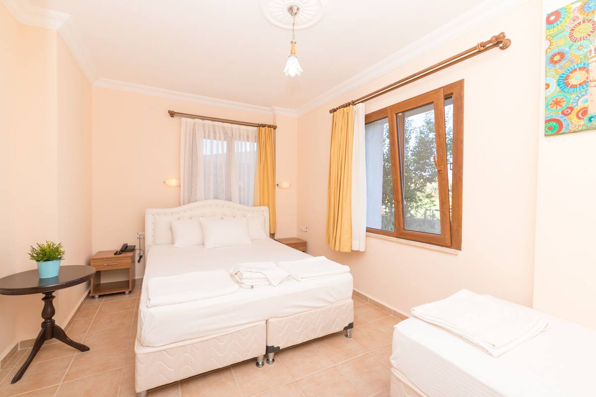A bedroom with a bed, bedside tables, a small table with a plant, a window with curtains, and a painting on the wall.