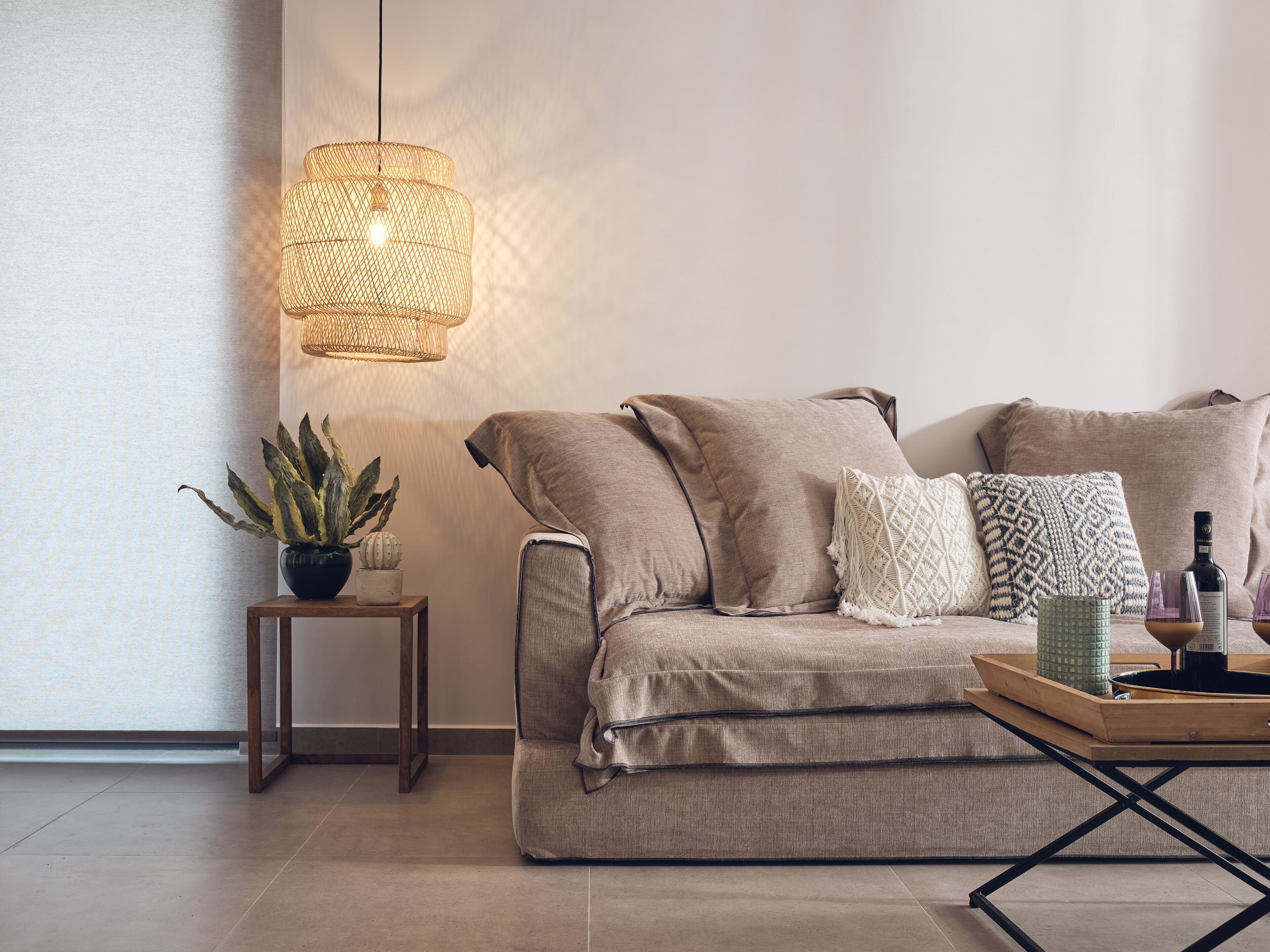 A modern living room with a beige sofa, a small wooden side table, a hanging light fixture, and a tray with a bottle and glasses.