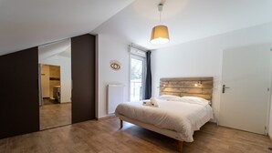 Un dormitorio con suelo de madera, una cama con ropa blanca, cabecero de madera, una ventana con cortinas y un espejo en la pared.