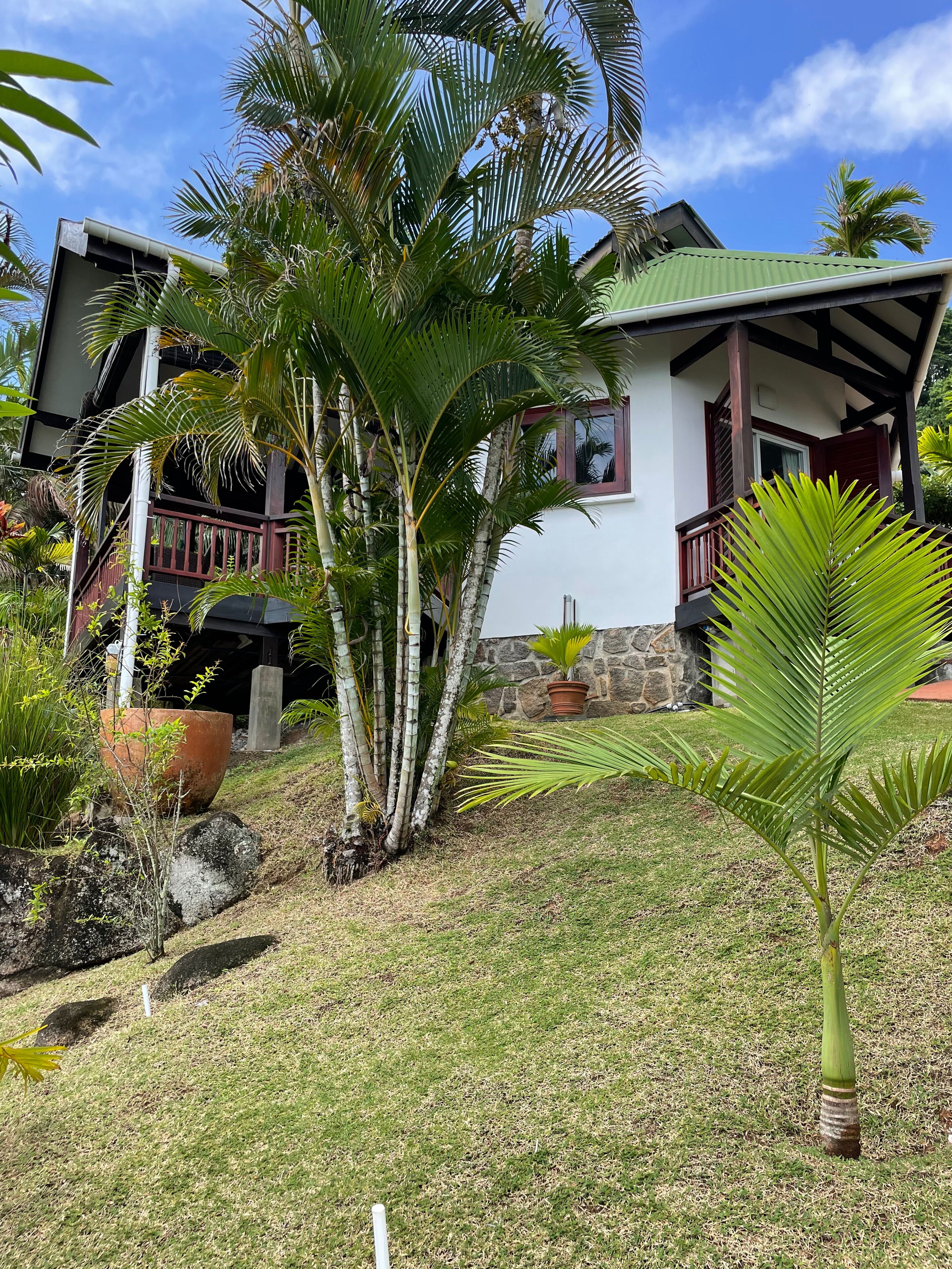 A two-story house with a balcony, surrounded by tropical vegetation and a stone wall.