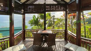 A wooden deck with a table and chairs, overlooking a tropical landscape with palm trees and the ocean. - Chalets Bougainville
One-Bedroom Chalet with Sea View (BOUGAINVILLE)