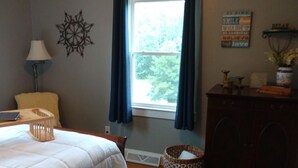 A bedroom with a bed, a nightstand, a lamp, a dresser, a window with curtains, a wall decoration, and a rug.