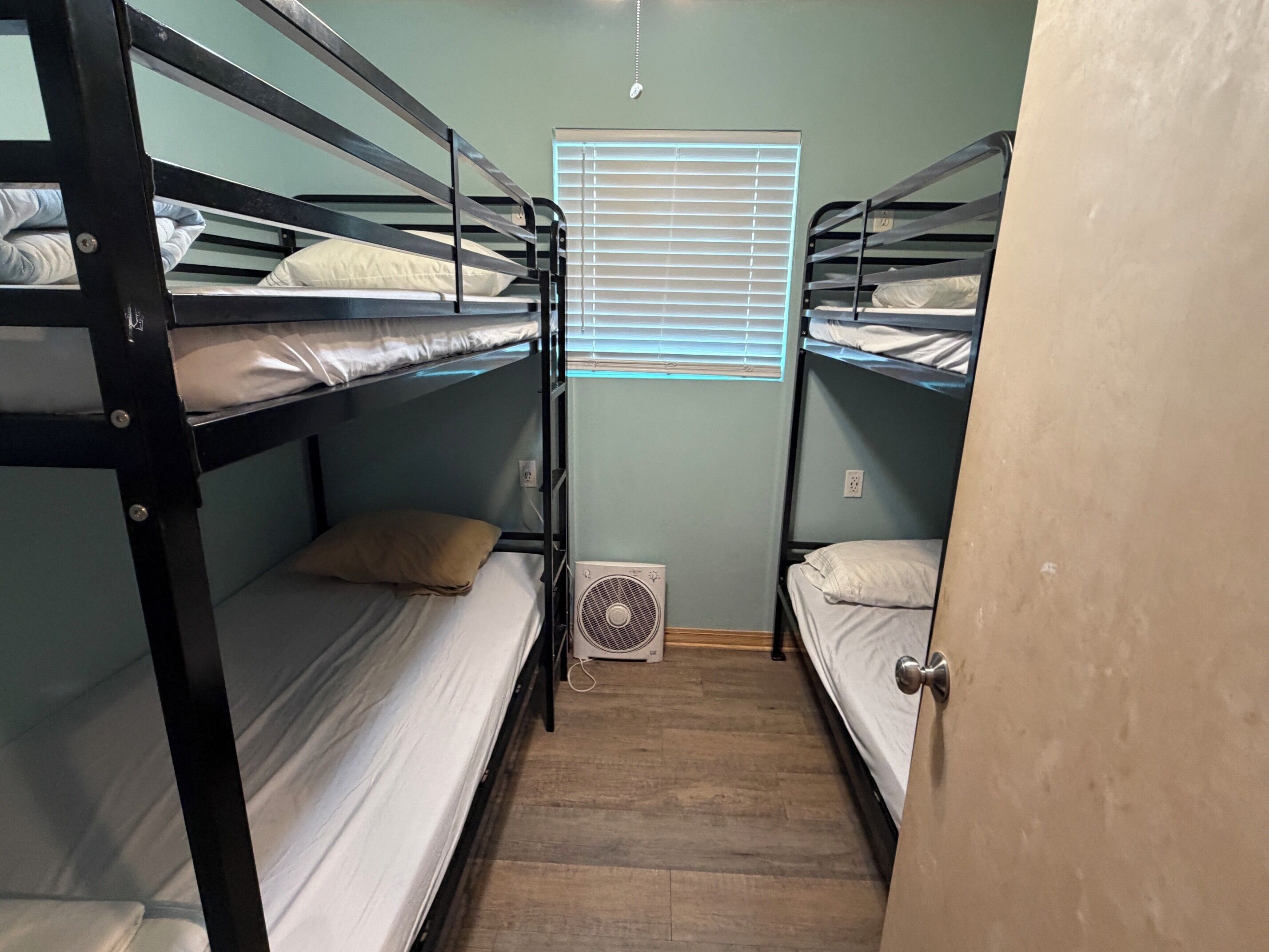 Une chambre avec deux lits superposés, une fenêtre avec des stores, un ventilateur fixé au mur et un plancher en bois.