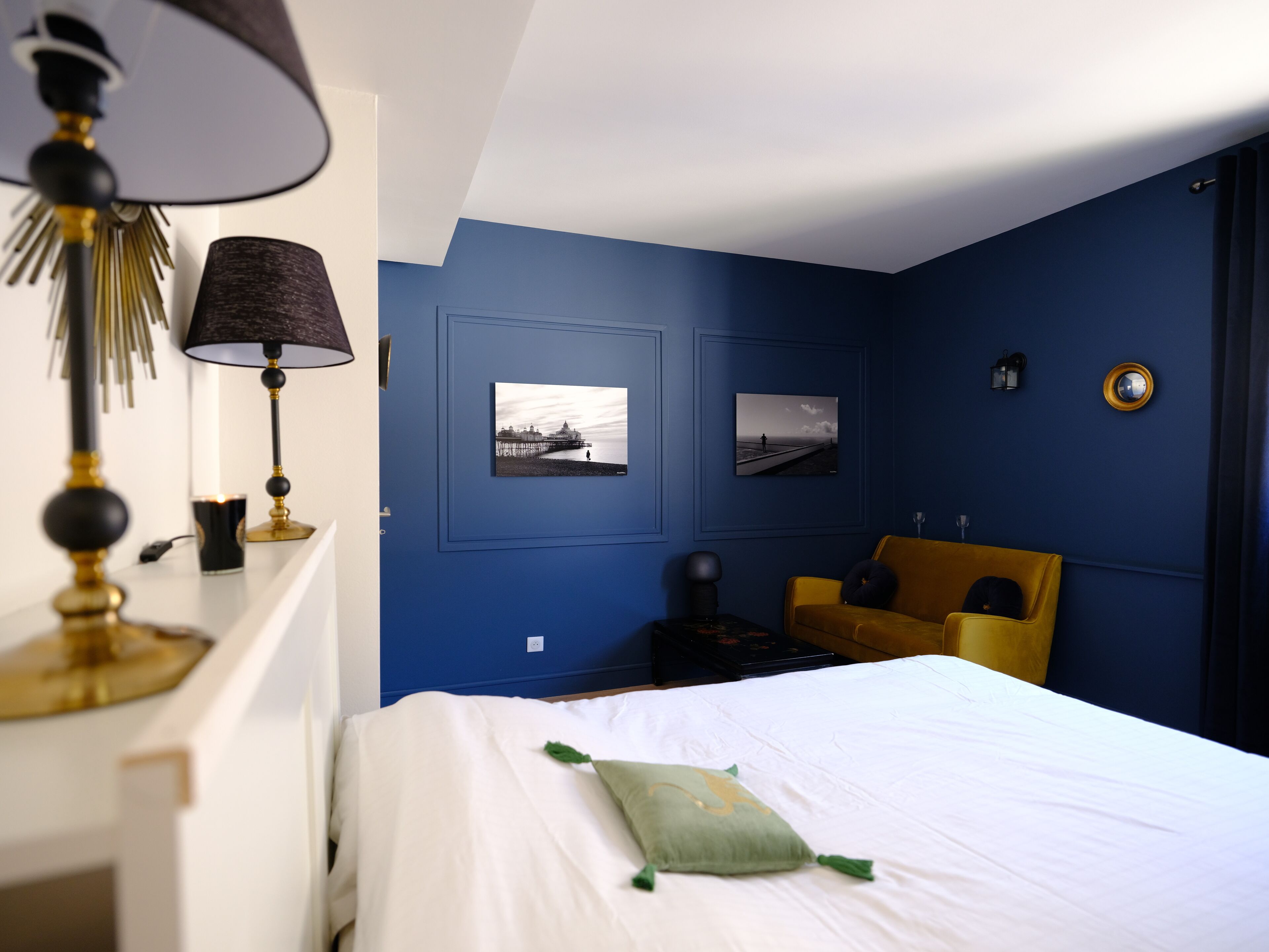Una habitación con una cama, un escritorio con lámpara, dos cuadros en la pared, una silla amarilla y una mesita con una vela.