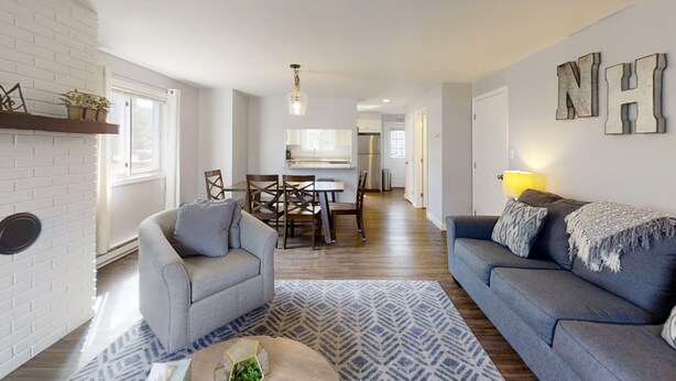 A modern living room with a grey sofa, a grey armchair, a dining table with chairs, and a kitchen in the background.