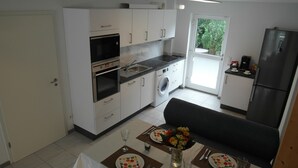 Cocina compacta con horno, microondas y lavadora incorporados. Cuenta con una zona de comedor con una mesa preparada para dos personas, y un jarrón con flores.
