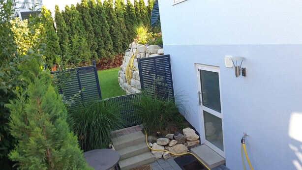 Un espacio exterior moderno con un muro de piedra, un pequeño jardín y un edificio azul.