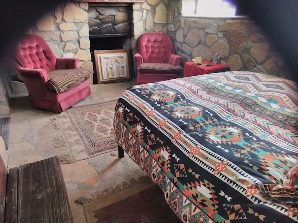 Une pièce avec une cheminée en pierre, deux fauteuils rouges, une couvre-lit à motifs et une petite table recouverte d’une nappe rouge.