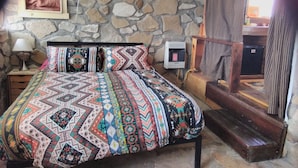 A room with a stone wall, a bed with patterned bedding, a bedside lamp, a small heater, and a wooden bench. - Stone cottage@ La loma del chivo (Marathon)