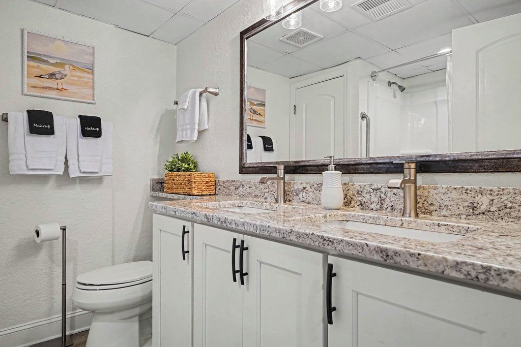 A bathroom with a white toilet, a sink with a marble countertop, a large mirror, and towels hanging on hooks.