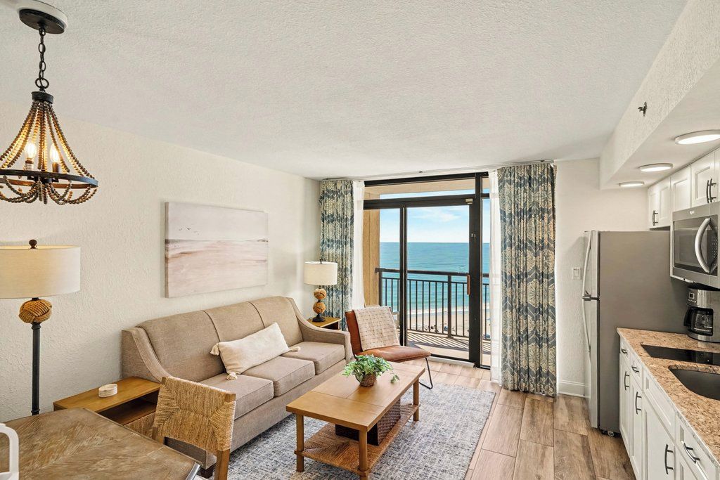 A modern living room with a sofa, armchairs, a coffee table, and a chandelier. There is a view of the ocean through a balcony door.