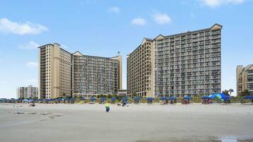 Un hotel frente a la playa con varios edificios altos, una playa de arena y sombrillas.