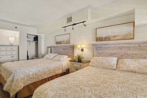 A hotel room with two beds, a dresser, a nightstand with a lamp, and a wall-mounted shelf.