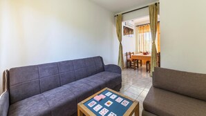 Uma sala de estar com um sofá cinzento, uma mesa de centro de madeira com cartas de baralho e uma área de jantar com mesa e cadeiras.